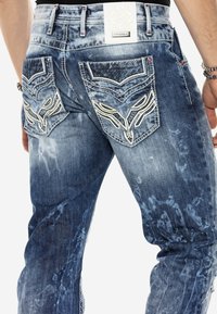 Herren Jeans aus blauen Denim mit einem beschädigten Finish, ausgestattet mit einzigartig bestickten Gesäßtaschen und einem geprägten weißen Lederpatch an der Taille.