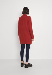 Langere rode jas met een kraag, gemaakt van zachte stof, met een rechte snit. Gecombineerd met donkerblauwe slim fit broek en zwarte schoenen.