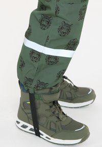 Groene waterdichte broek met geometrische tijgerpatronen en reflecterende strepen, gecombineerd met olijfgroene hoge sneakers met textuuraccenten.