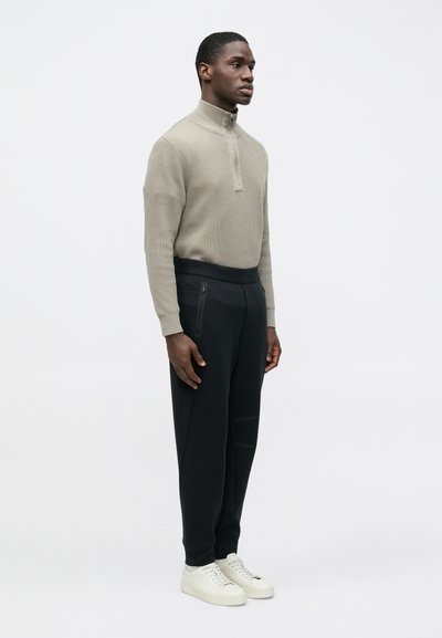 Μπεζ πουλόβερ με ραβδώσεις και μισό φερμουάρ, σε συνδυασμό με μαύρο παντελόνι με κωνική γραμμή. Το παντελόνι διαθέτει δύο πλευρικά φερμουάρ. Το μοντέλο φορά λευκά αθλητικά παπούτσια.