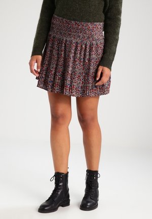 Kvinde iført en grøn sweater, plisseret blomsterprintet miniskørt og sorte snørestøvler, stående på en hvid baggrund.