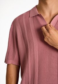 Pletené polo tričko v jemné růžové barvě s límečkem. Disponuje vertikálním žebrováním a krátkými rukávy. Hladká, měkká látka.