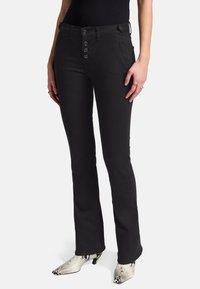 Jean noir évasé avec fermeture à cinq boutons, accents en métal, poches avant et un tissu en coton mélangé élégant. Porté avec des bottes à motifs à la cheville.