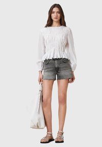 Blouse blanche à manches longues avec un design plissé et un ourlet peplum, associée à un short en denim gris. Le modèle tient un grand sac fourre-tout blanc. Sandales argentées.
