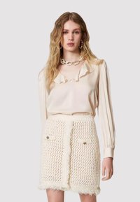 Blusa crema con collo a volant, abbinata a una gonna crema con texture e dettagli di bottoni e frange. Tessuto liscio e design intricato.