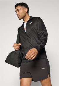 Sort zip-up jakke med Puma-logo, parret med grå shorts med sidelæg. Modellen bærer en sort duffelbag, hvilket fremhæver et sporty look.
