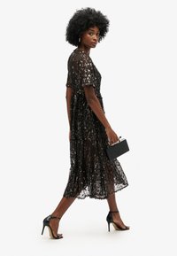 Vestido negro con lentejuelas, de manga corta, hasta la rodilla, con detalles de flecos. Combinado con sandalias negras de tacón alto y un pequeño bolso de mano cuadrado.