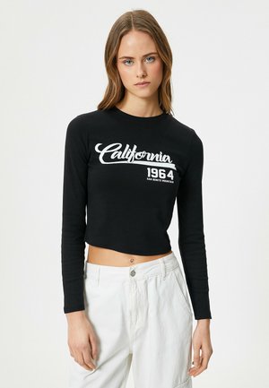 Zwarte cropped langemouwen t-shirt met witte tekst "California 1964" in gebogen letters; gecombineerd met losse witte broeken.