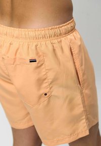 Lichtperzik kleurige zwembroek met een elastische tailleband, zijzakken en een achterzak met een zwart, rood en wit streepdessin.