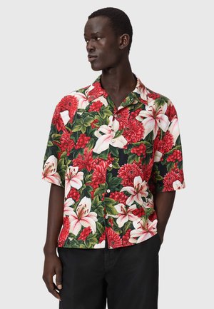 Mand iført kortærmet skjorte med knapper og dristigt rødt og hvidt blomsterprint, stående med den ene hånd i lommen og kigger til venstre.