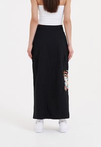 Sort maxi-nederdel med to side lommer, prydet med farverig broderi af et ansigt på højre side. Lavet af et glat stof.