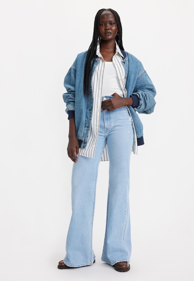 Jeansjacke mit oversize Passform und blauer Farbe, geschichtet über einem weißen, gestreiften Hemd, kombiniert mit hellblauen Schlagjeans. Braune Schuhe.