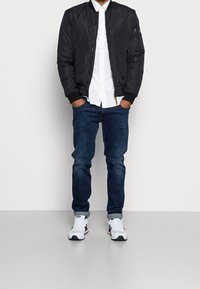 Svart bomberjacka, vit skjorta med knapp, mörka jeans och vita sneakers. Jackan har ribbade mudd; jeansen har en smal passform med en subtil blekning.