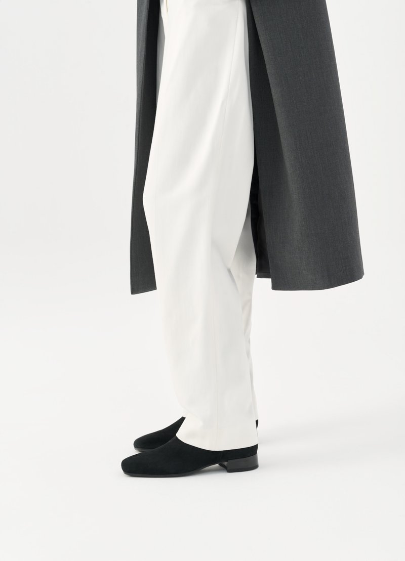 Schwarze Wildlederschuhe, weiße maßgeschneiderte Hosen und ein dunkelgrauer langer Mantel, die ein schlankes und elegantes Aussehen betonen.