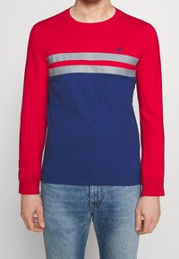 Maglietta a maniche lunghe rossa e blu, con strisce orizzontali grigie sul petto. Realizzata in cotone, con collo a giro e vestibilità regolare.