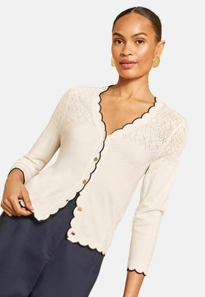 Frau mit nach hinten gekämmtem Haar trägt einen cremefarbenen Cardigan mit schwarzen, gewellten Kanten und goldenen Knöpfen, kombiniert mit einer dunklen Hose und goldenen Ohrringen.