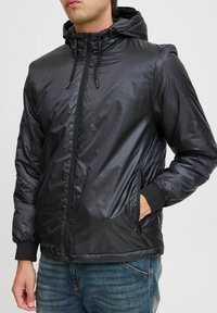 Schwarze, leichte Kapuzenjacke mit einer glatten, glänzenden Textur. Verfügt über einen durchgehenden Reißverschluss, elastische Bündchen und zwei Seitentaschen.