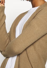 Person som bär en strukturerad beige stickad cardigan över en vit t-shirt, med en arm lyft och delvis böjd.