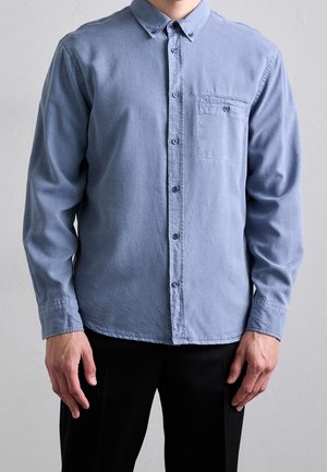 Homme portant une chemise bleu clair à manches longues avec une poche poitrine, associée à un pantalon noir, debout devant un fond uni.