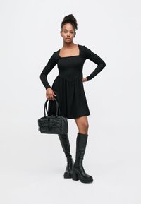 Zwarte geribbelde jurk met lange mouwen en een vierkante halslijn, gecombineerd met zwarte plateau laarzen tot aan de knie en een kleine zwarte handtas met strikdetails.