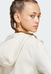 Un sweat à capuche couleur crème avec une texture douce, doté d'une capuche à cordon et de détails cousus, porté par une personne avec des cheveux tressés.