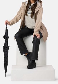 Femme portant un imperméable beige, un pull gris, un pantalon noir et des bottes noires assise sur un bloc blanc tenant un parapluie noir fermé.