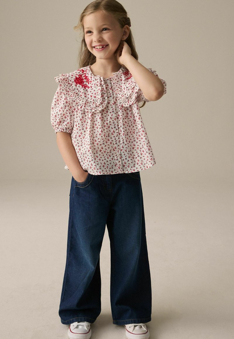 Weiße Bluse mit kleinem roten Blumenmuster und Rüschenkragen, kombiniert mit weit geschnittenen blauen Jeans und weißen Sneakern.