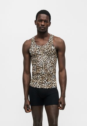 Jeune homme à la peau foncée portant un débardeur sans manches imprimé léopard et un short noir moulant, debout devant un fond clair uni.