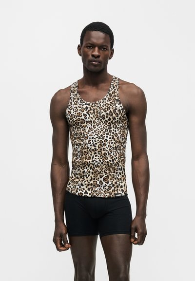Jeune homme à la peau foncée portant un débardeur sans manches imprimé léopard et un short noir moulant, debout devant un fond clair uni.