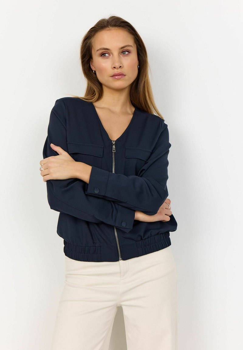 Marineblauwe cropped jas met een rits aan de voorkant, twee borstzakken en een elastische tailleband; gemaakt van een gladde stof met een lichte glans.