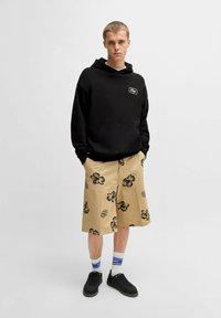 Felpa nera con cappuccio e tasca frontale, pantaloni beige con motivi floreali neri, sneaker nere e calzini bianchi fino alla caviglia con dettagli blu.