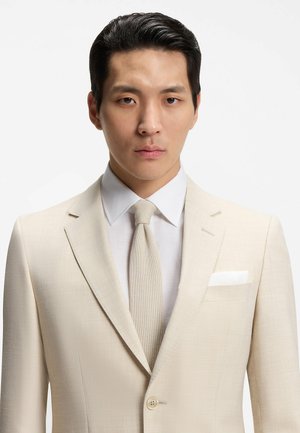 Junger Mann mit schwarzen Haaren, der einen beigen Anzug, ein weißes Hemd und eine beigefarbene Strickkrawatte trägt, frontal vor einem schlichten weißen Hintergrund.