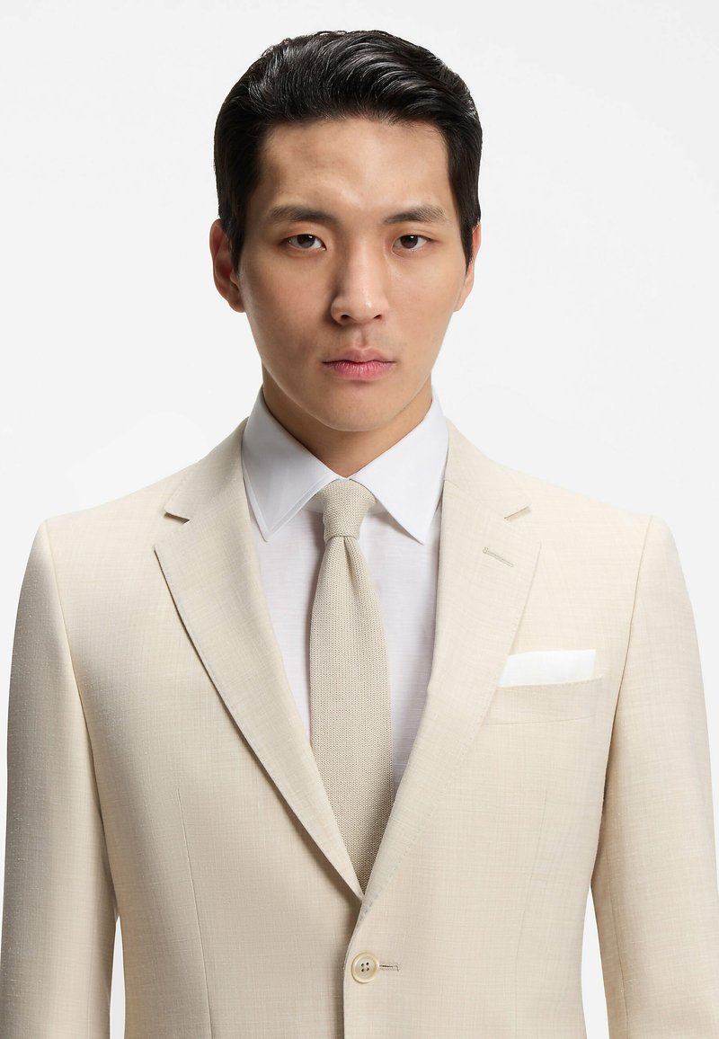 Jeune homme aux cheveux noirs portant un costume beige, une chemise blanche et une cravate en maille beige, faisant face à l'avant sur un fond blanc uni.