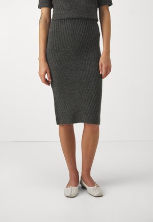 Femme portant une jupe crayon tricotée texturée gris foncé longueur genoux et des ballerines blanches, debout sur un fond blanc uni.