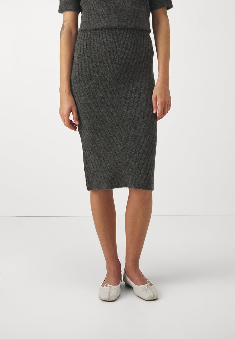 Femme portant une jupe crayon tricotée texturée gris foncé longueur genoux et des ballerines blanches, debout sur un fond blanc uni.