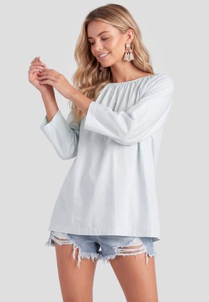 Junge Frau mit langen blonden Haaren trägt ein weißes langärmeliges Oberteil, zerrissene Denim-Shorts und große Quastenohrringe und richtet ihre Hand.