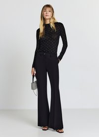 Top nero a maniche lunghe con pois bianchi, abbinato a pantaloni neri a vita alta e svasati. La modella tiene in mano una piccola borsa metallica.