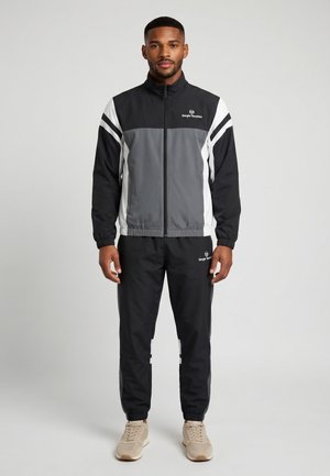 Hombre de pie mirando hacia delante, viste un chándal Sergio Tacchini en negro, gris y blanco con zapatillas beige sobre un fondo liso.