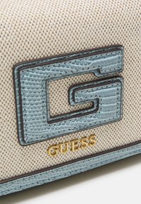 Beige textilní plátěná kabelka s modrým koženým logem "G" a švy, pod kterým je zlaté značení "GUESS".