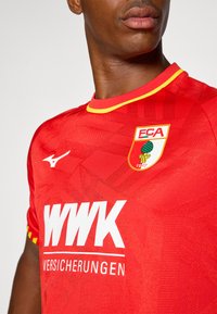 Sarkans futbols krekls ar ģeometrisku rakstu, dzelteniem akcentiem un apaļu apkakli. Iekļauto logotipu: "FCA" un "WWK Versicherungen" balti.