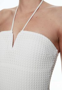Haut blanc texturé avec un décolleté en V, présentant un motif tissé et des fines bretelles, mettant en avant une coupe ajustée.