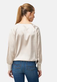 Hedvábná halenka světle béžové barvy s dlouhými, volánovitými rukávy. Zadní část má zaoblený výstřih, doplněná přiléhavými modrými džíny s detailními kapsami.