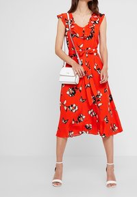 Robe fleurie rouge avec un col à volants et des manches courtes, dotée d'une taille cintrée par une ceinture et d'une jupe fluide. Associée à un sac à main blanc et des sandales.