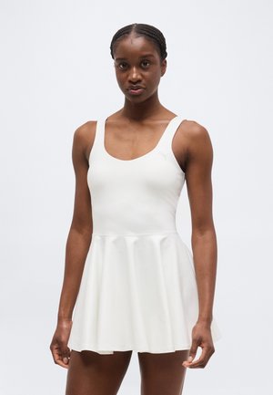 Naine, kes kannab valget varrukatut tennisekleiti koos keha järgiva ülakeha ja lehviva seelikuga, seisab ühevärvilise heleda tausta ees.