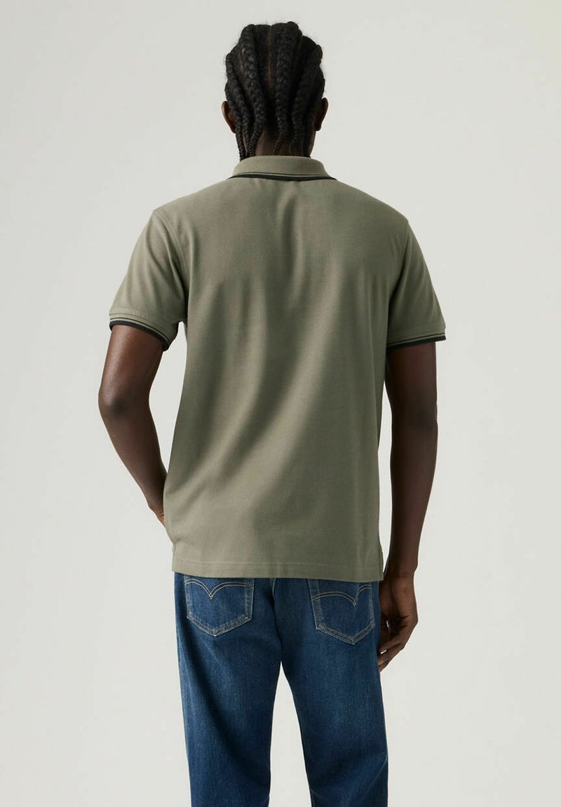 Polo verde oliva con maniche corte e rifinitura nera a contrasto sul colletto. Indossata con jeans di denim blu. La texture appare liscia.