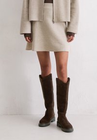 Frau trägt beigen Rock, beige Jacke mit Reißverschluss und dunkelbraune kniehohe Wildlederstiefel, steht vor einer hellen Wand.