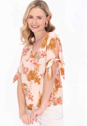 Femeie zâmbitoare cu păr blond ondulat, purtând o bluză cu flori cu mâneci scurte și fundițe legate și pantaloni albi pe un fundal alb simplu.