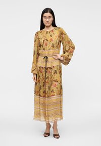 Μακρύ φόρεμα με σχέδιο σε μουσταρδί κίτρινο χρώμα με λουλουδάτα μοτίβα και σφιχτή μέση. Διαθέτει μακριά μανίκια και φούστα με βολάν. Καφέ παπούτσια.