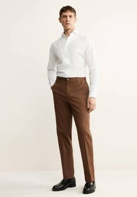 Chemise blanche à boutons, pantalon marron sur mesure, chaussures en cuir noires. Tissu lisse, design ajusté, et détails de couture subtils sur le pantalon.