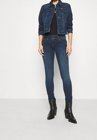 Mujer con chaqueta de mezclilla oscura y jeans, camiseta negra y botas de tacón negro hasta el tobillo, de pie contra un fondo claro y liso.